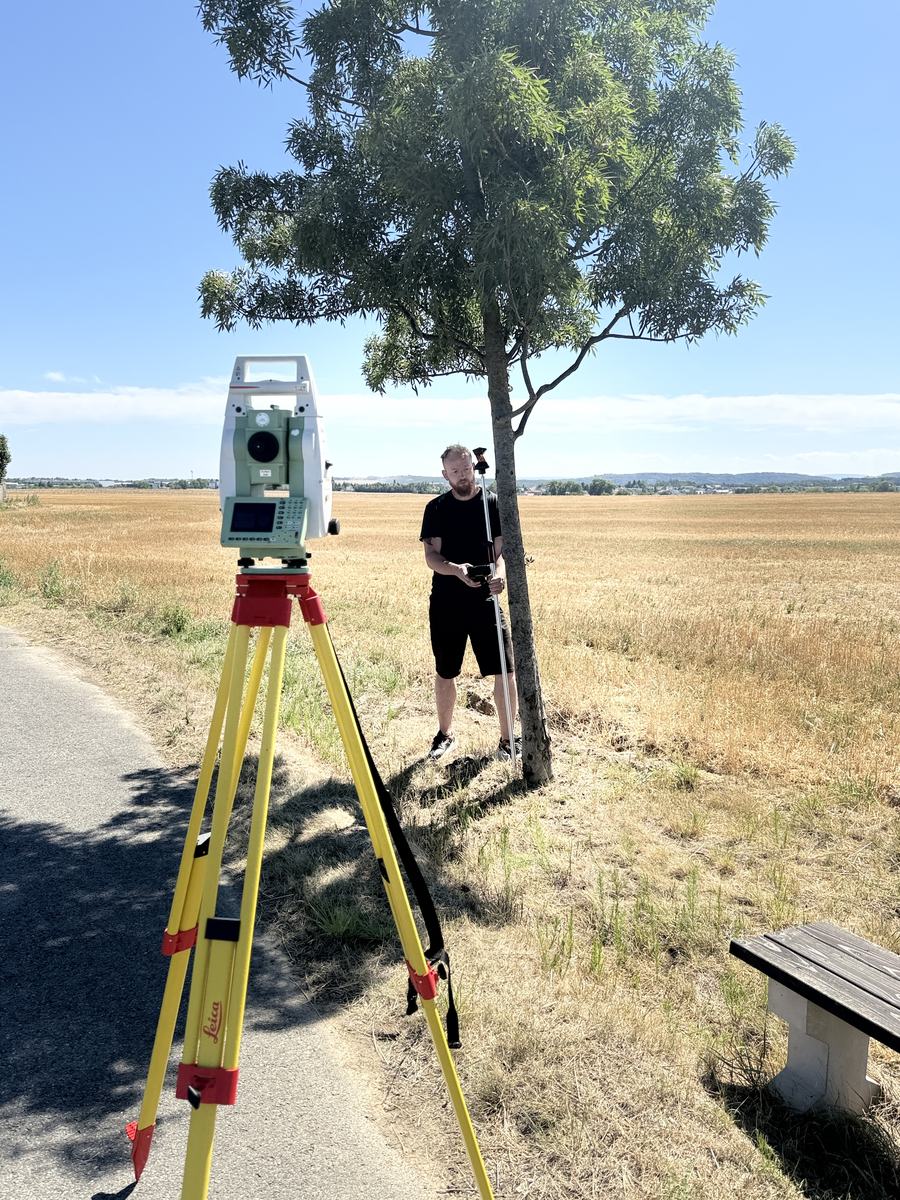 Robotická totální stanice Leica TCRP 1203 na stativu u cesty, v pozadí geodet INGsite s výtyčkou pod stromem na okraji pole